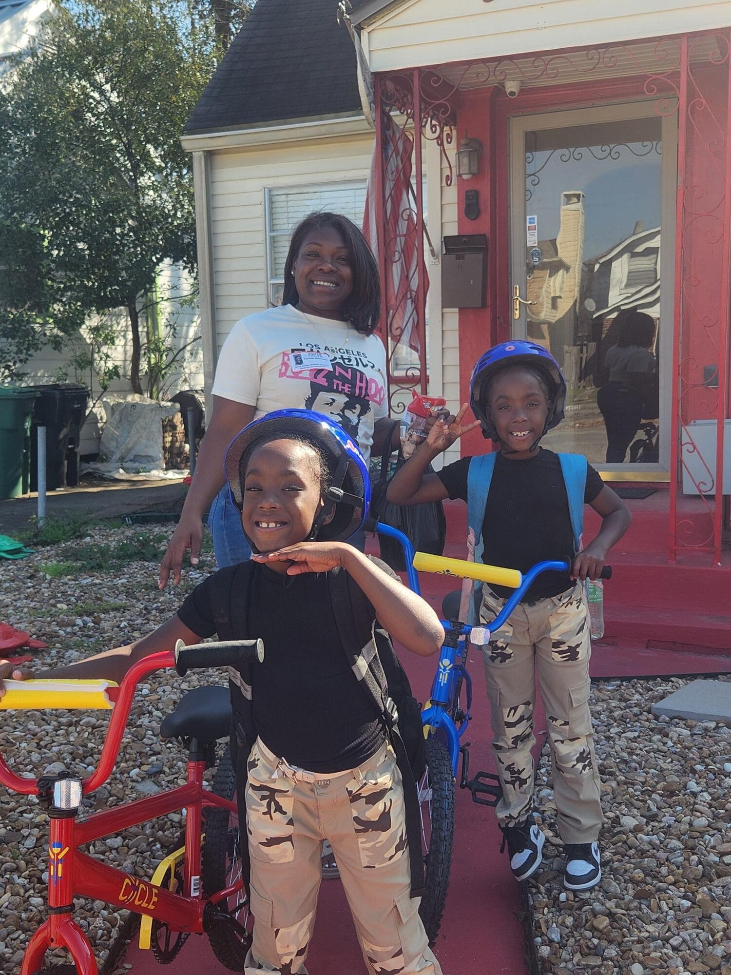 Two kids with bikes and helmets pose with a woman in front of a red house.